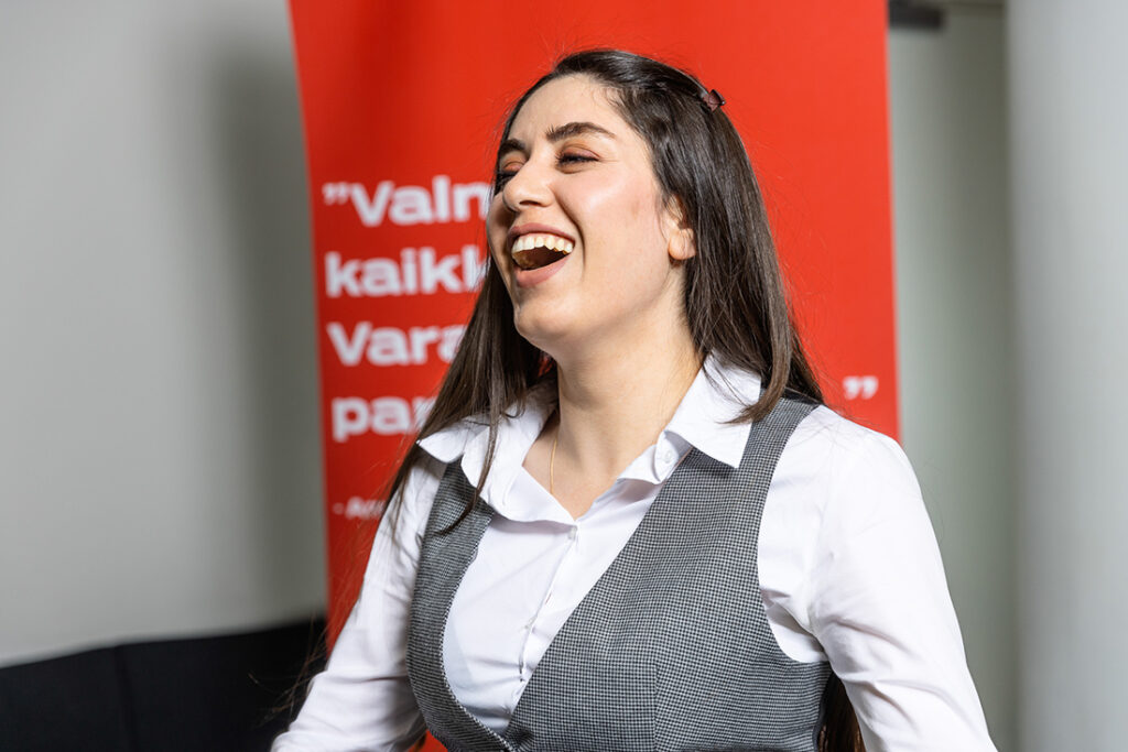 Nuori naisoletettu nauraa jollekin. Taustalla näkyy TAIn punainen rollup.
