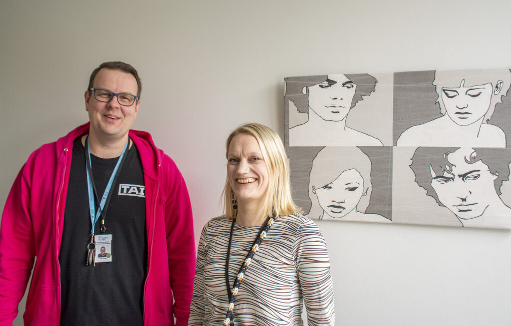 TUVA-ohjaaja Kalle Kaskeala ja opinto-ohjaaja Kaisu Kippo poseeraavat kameralle vaalean seinän edessä. Taustalla näkyy grafiikka tms. taideteos, jossa on neljä piirrettyä ihmishahmoa.