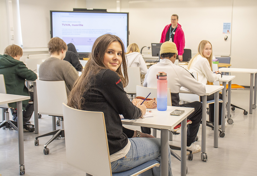 A student looks at a camera in a small classroom. In the background, you can see the backs of other students and the teacher in front of the class.